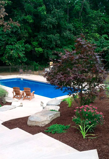 A patio adjacent to a pool featuring outdoor fireplace, Adirondack chairs and attractive landscape featuring boulders and decorative shrubs