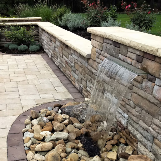 A decorative stone masonry retaining wall filled with plants and shrubs featuring a waterfall and multi colored stone patio