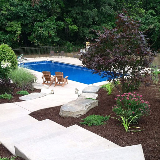 A patio adjacent to a pool featuring outdoor fireplace, Adirondack chairs and attractive landscape featuring boulders and decorative shrubs