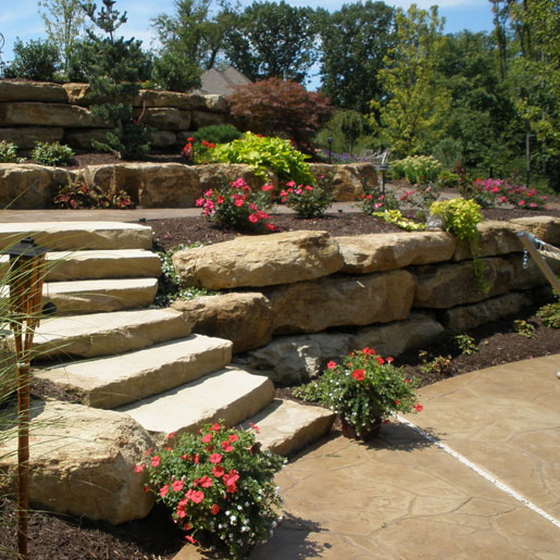 A set of retaining walls made of boulders with stone steps and planter boxes filled with dirt and flowering annual plants